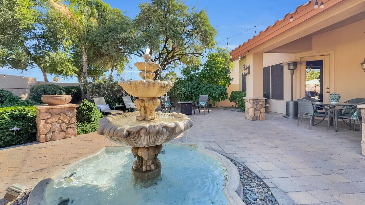 A serene exterior scene featuring a bubbling fountain, comfortable patio seating, and lush greenery surrounding the area. The space suggests a peaceful ambiance perfect for relaxation or socializing outdoors.