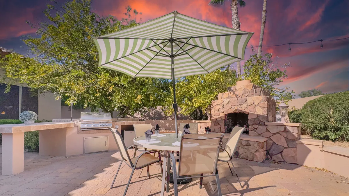 A spacious backyard patio features a table under an umbrella, a cozy fireplace, a grill, and ample greenery in the background. The scene invites both intimate fireside chats and festive barbecues, emphasizing the space's versatility.
