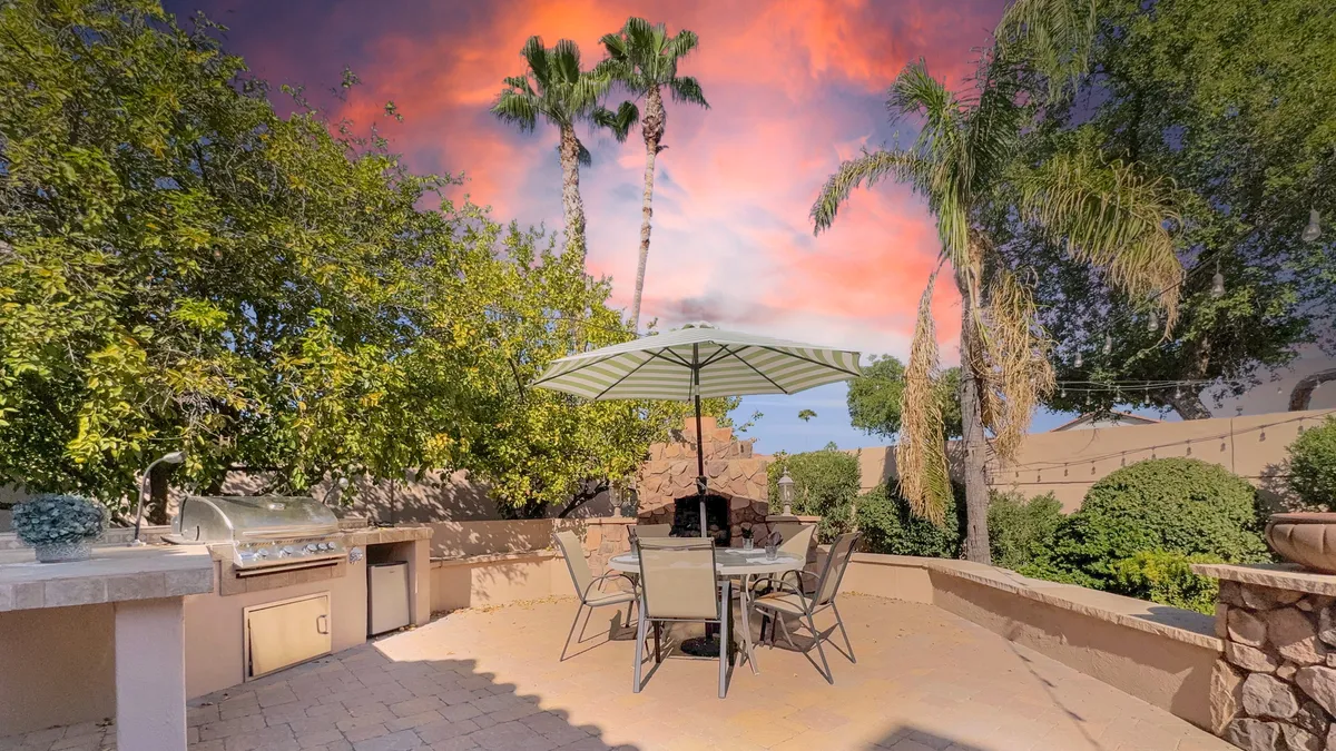 A relaxed outdoor dining set for eight sits beneath a large umbrella, flanked by a modern grill and shaded by palm trees. The area is surrounded by lush greenery, creating a private atmosphere ideal for al fresco meals and gathering with friends or family.