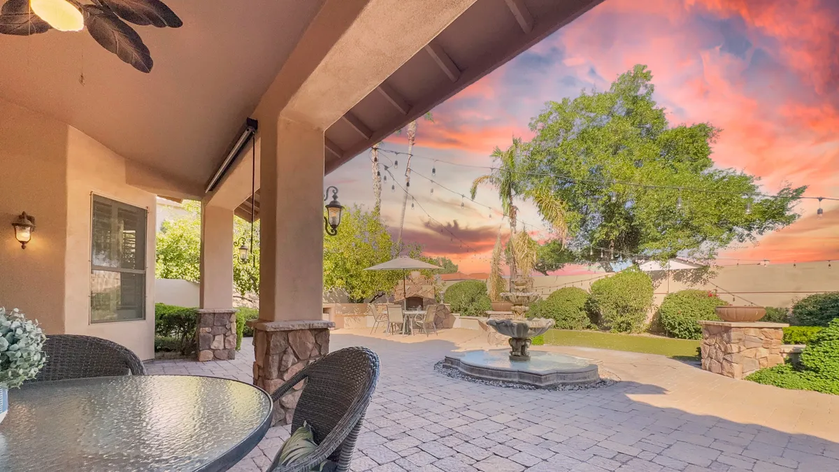 An inviting patio setting with a graceful fountain as its centerpiece, surrounded by lush landscaping and set against a colorful sunset sky. The space is well-appointed for outdoor dining, social gatherings, or quiet moments.