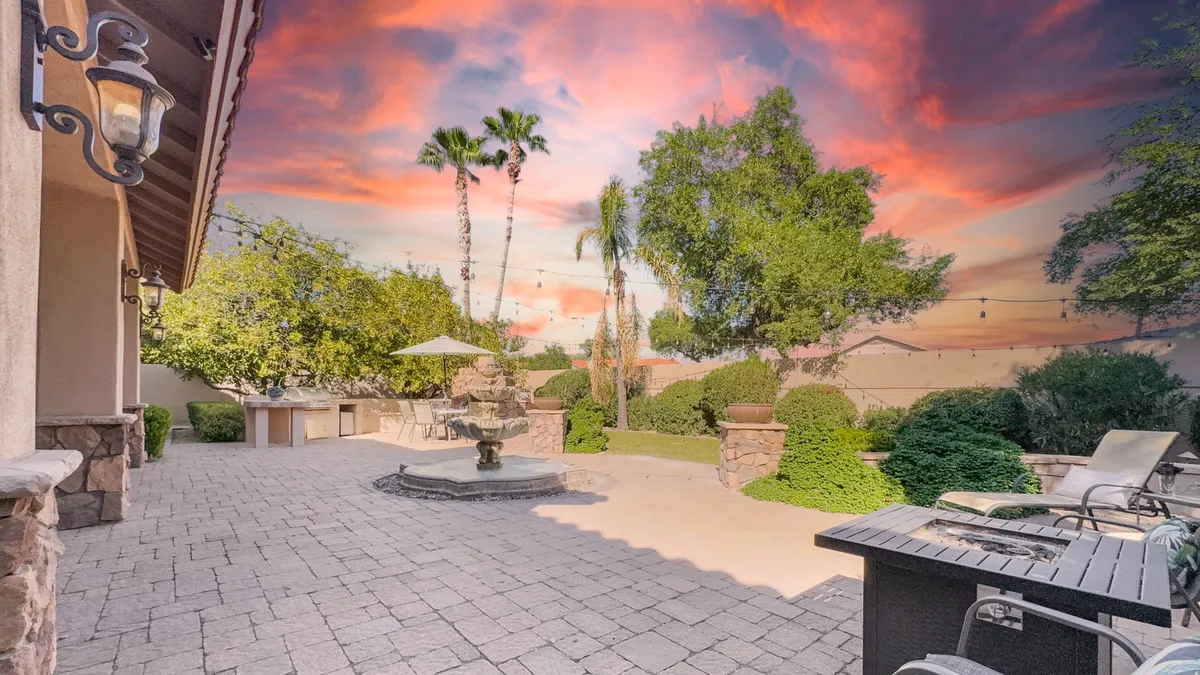 An inviting backyard scene with patio seating arranged for group gatherings, a bubbling fountain as a focal point, lush plantings, and the warm glow of a sunset in the background. The space is designed for outdoor dining, celebrations, and peaceful relaxation.